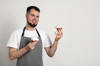 Photo of Happy professional confectioner in apron holding delicious cupcakes on light grey background. Space for text