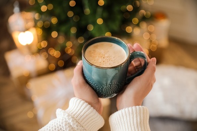 Photo of Woman holding cup of cocoa near Christmas tree at home, closeup