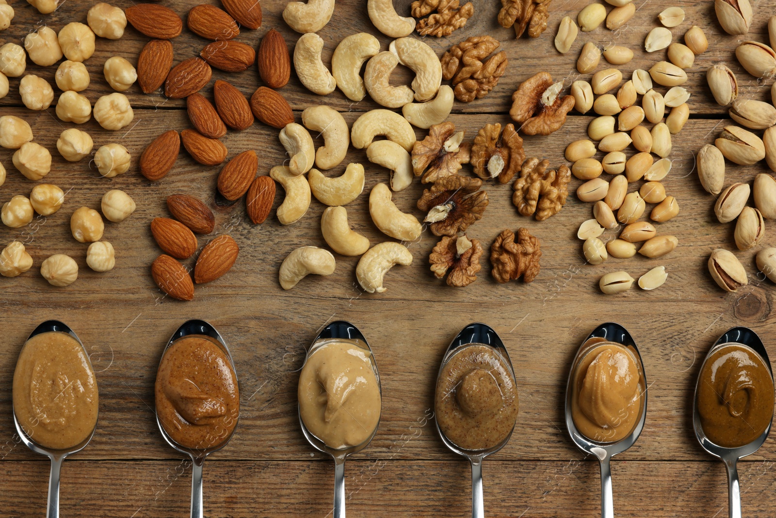 Photo of Tasty nut butters in spoons and raw nuts on wooden table, flat lay