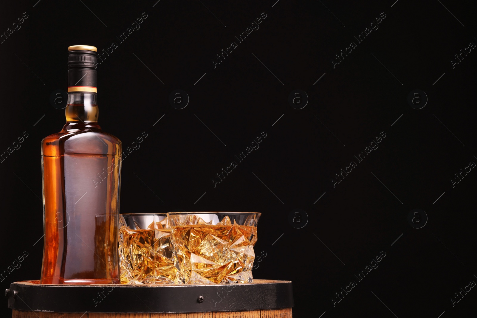 Photo of Whiskey in glasses and bottle on wooden barrel against black background, space for text