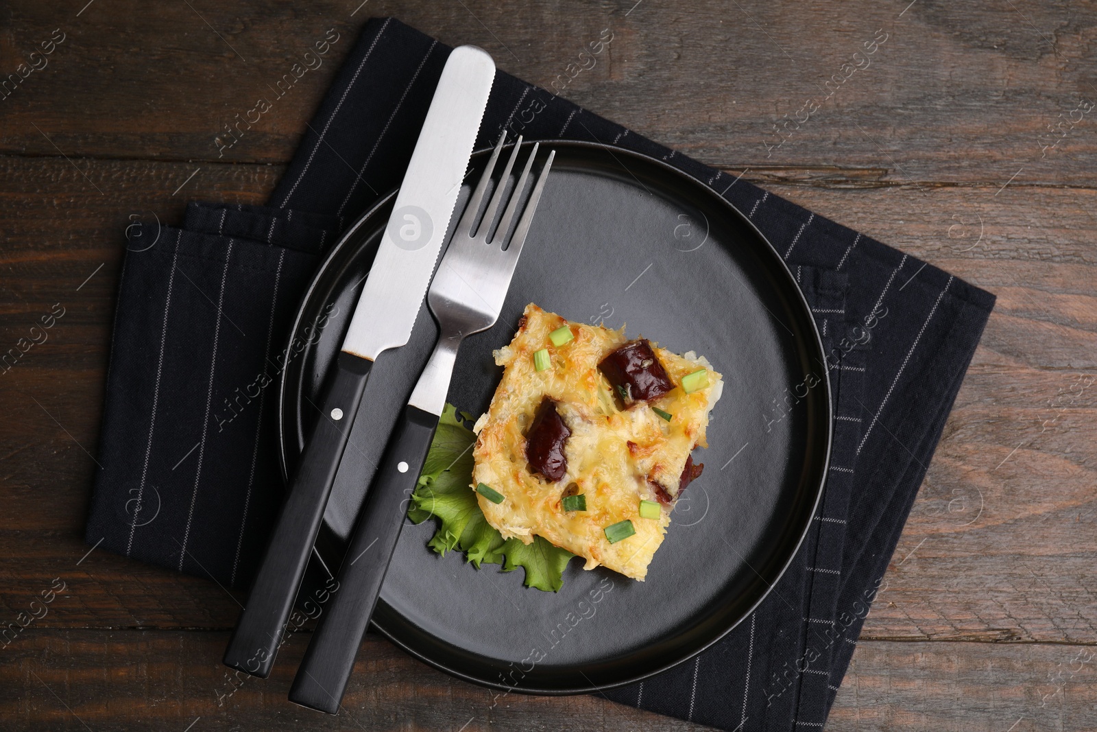 Photo of Tasty sausage casserole with green onions and cutlery served on wooden table, top view