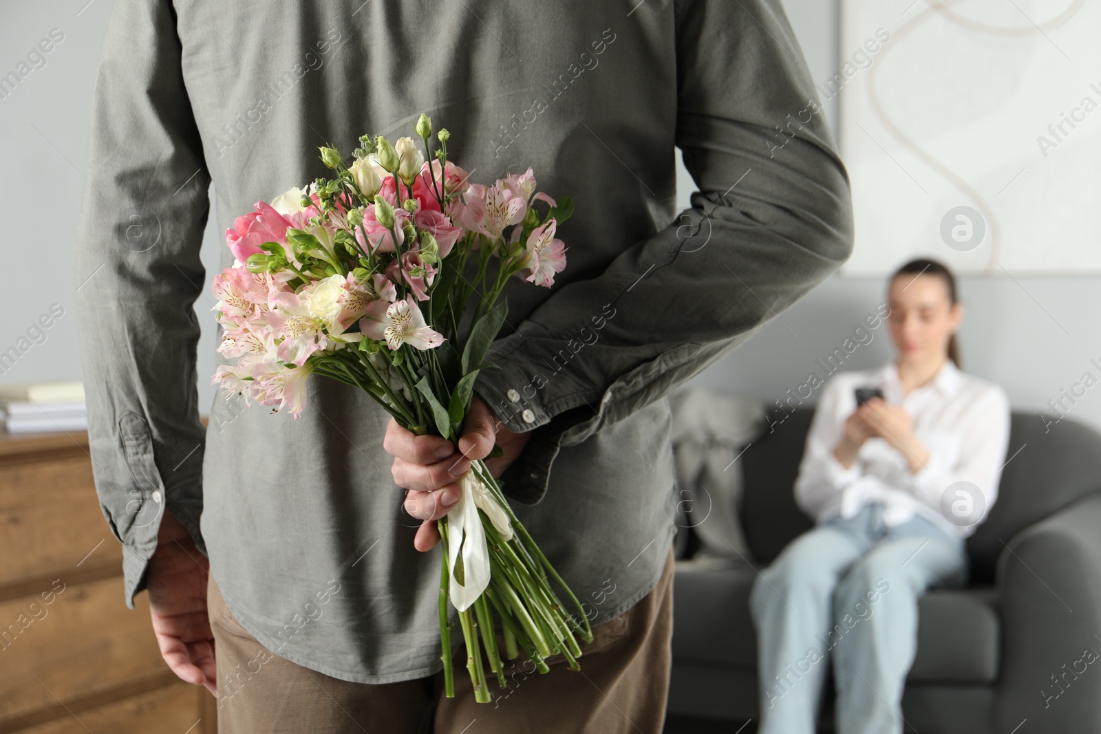 Photo of Man hiding bouquet of flowers for his beloved woman indoors, closeup