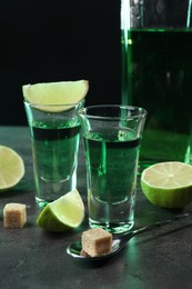 Photo of Absinthe in shot glasses, spoon, brown sugar cubes and lime on gray textured table, closeup. Alcoholic drink