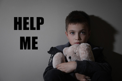 Image of Sad little boy with teddy bear and text HELP ME on beige background