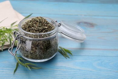 Photo of Jar of dry tarragon and green leaves on light blue wooden table, closeup. Space for text