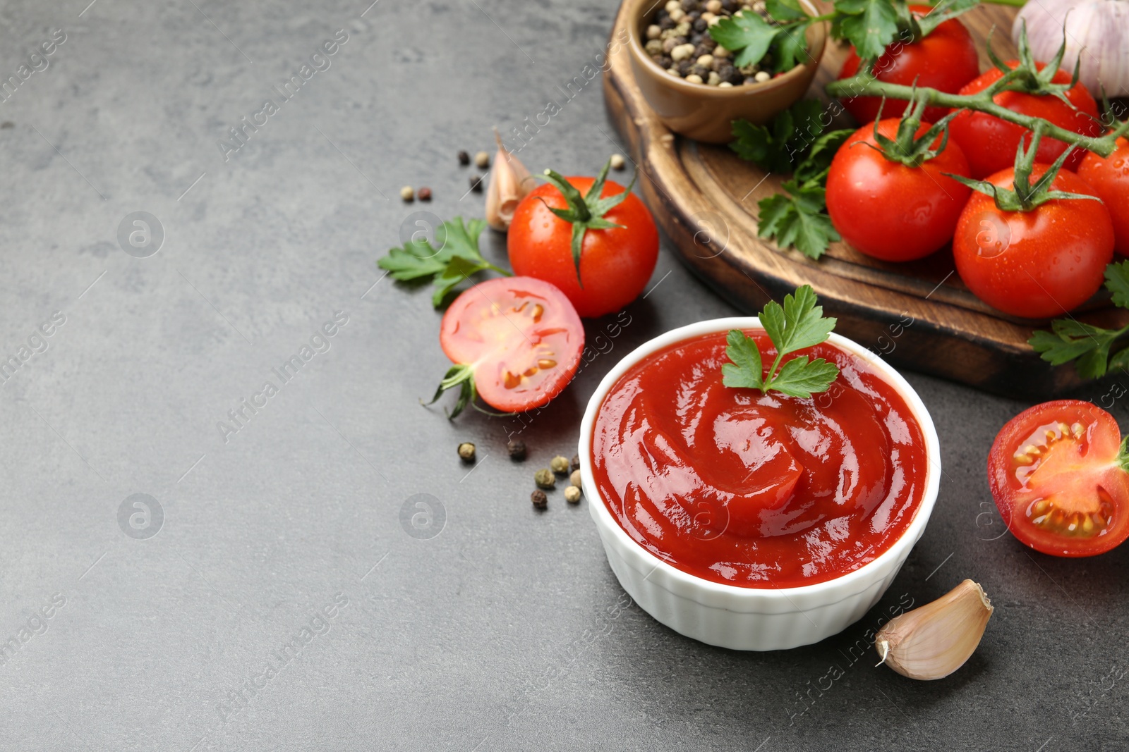 Photo of Delicious ketchup in bowl, tomatoes, parsley and garlic on grey table, closeup. Space for text