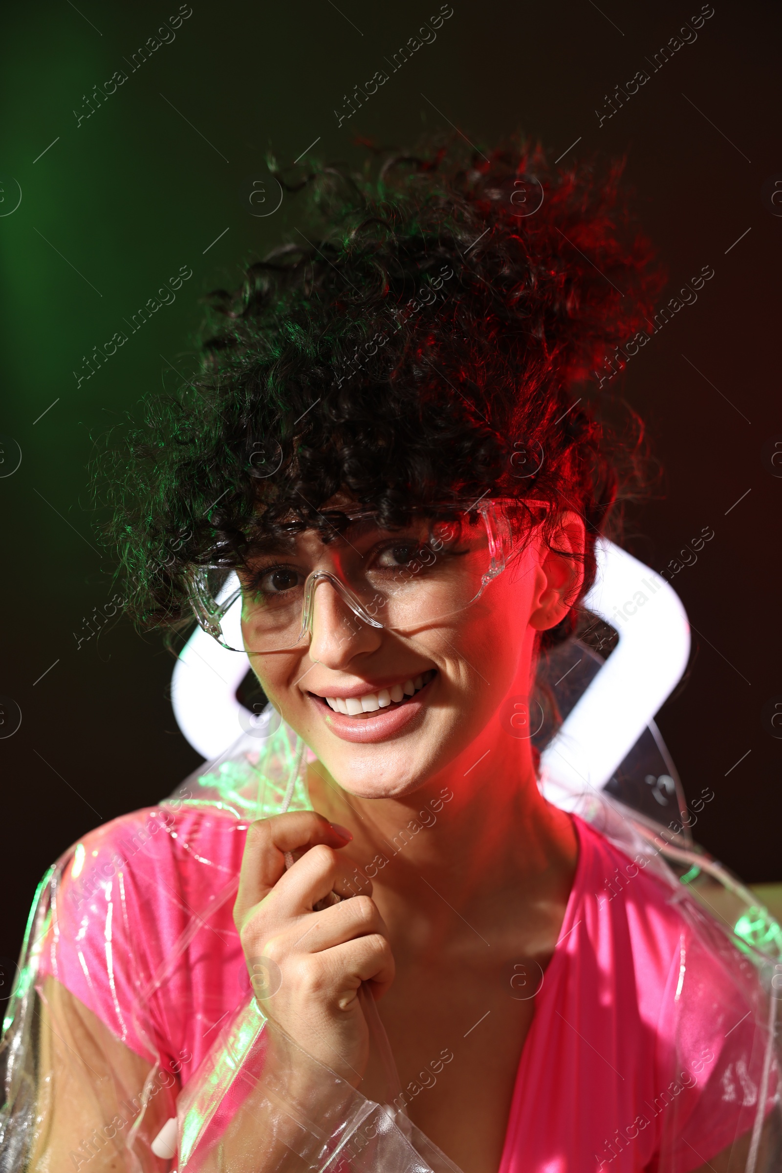 Photo of Beautiful young woman in transparent coat and sunglasses with square lamp on color background in neon lights