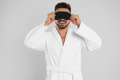 Young man in bathrobe and eye sleeping mask on light grey background