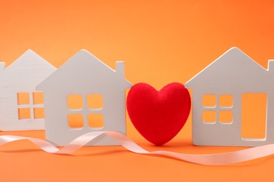 Photo of Long-distance relationship concept. Decorative heart between white house models and ribbon on orange background