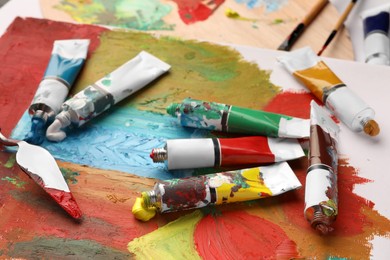 Photo of Tubes of colorful oil paints, spatula and canvas with abstract painting on table