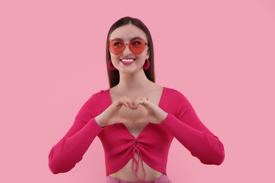 Photo of Pink look. Beautiful woman in sunglasses making heart with hands on color background
