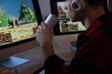 Young man with energy drink and headphones playing video game at wooden desk indoors, closeup