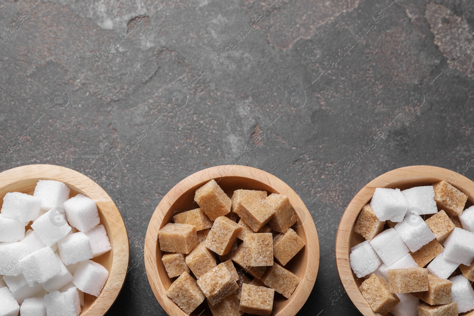 Photo of Different sugar cubes in bowls on gray textured table, flat lay. Space for text