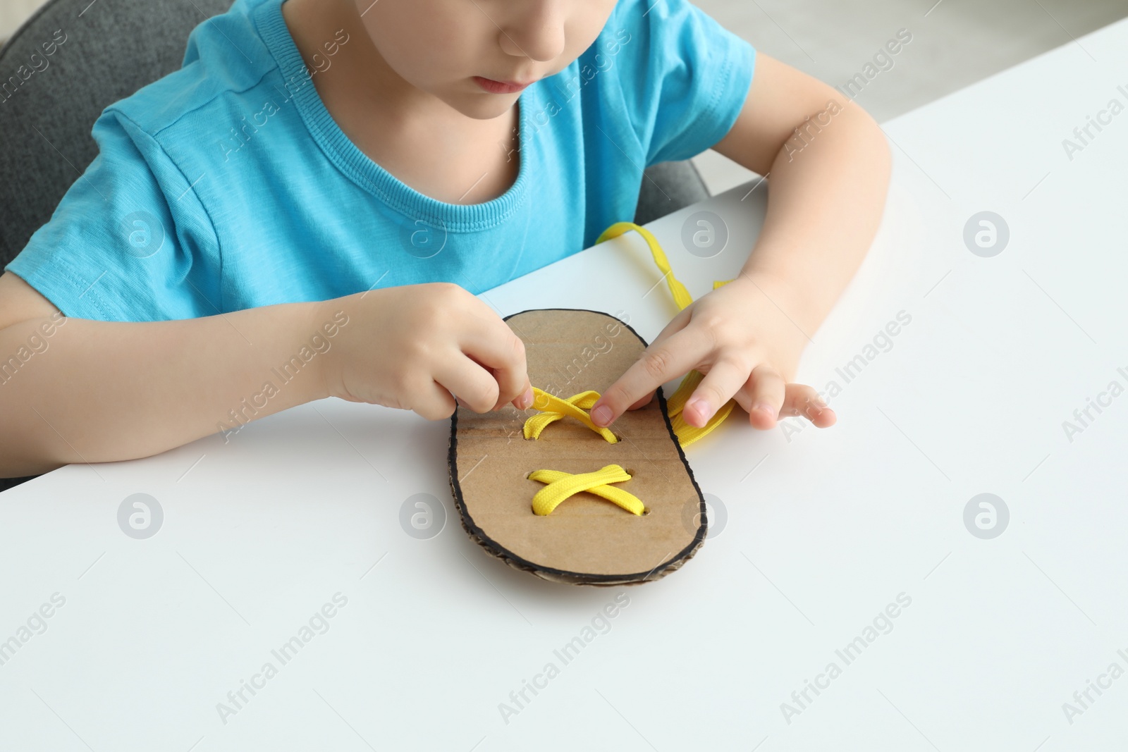 Photo of Little boy tying shoe lace using training cardboard template at white table, closeup