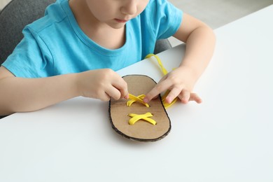 Little boy tying shoe lace using training cardboard template at white table, closeup