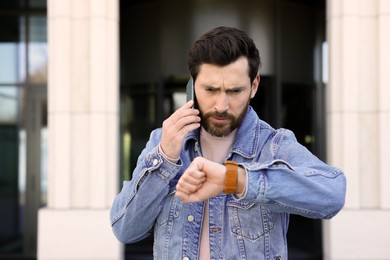 Photo of Unhappy man talking on smartphone and looking at watch near building. Being late concept