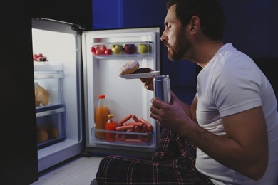 Man with donuts and drink near refrigerator in kitchen at night. Bad habit
