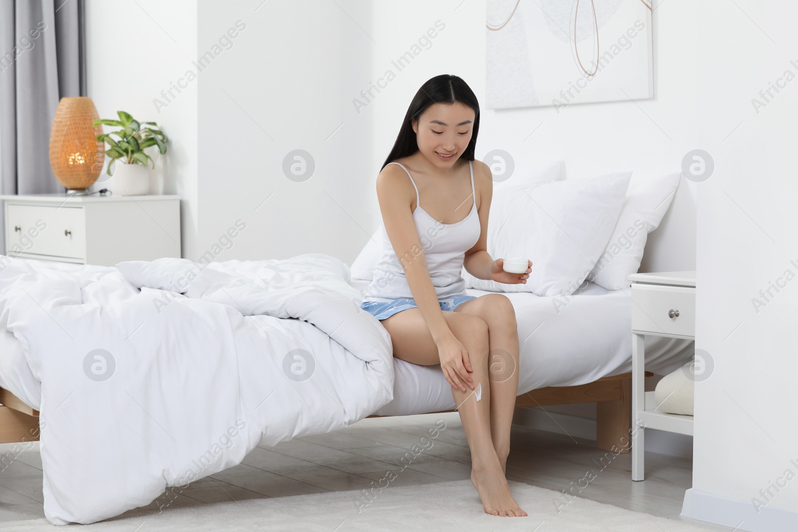 Photo of Beautiful young Asian woman applying body cream on leg in bedroom