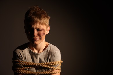 Photo of Little boy with bruises tied up and taken hostage on dark background. Space for text