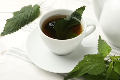 Photo of Cup of aromatic nettle tea and green leaves on white wooden table, closeup