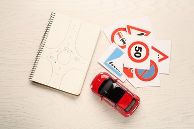 Photo of Many different road sign cards, notebook with sketch of roundabout and toy car on white wooden table, flat lay. Driving school