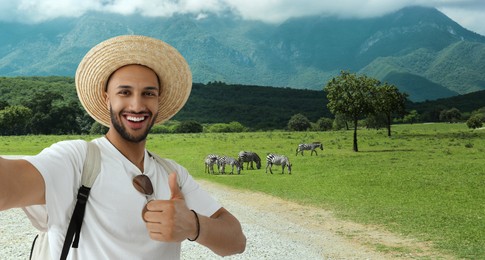Image of Smiling young man in straw hat taking selfie and showing thumbs up at nature park