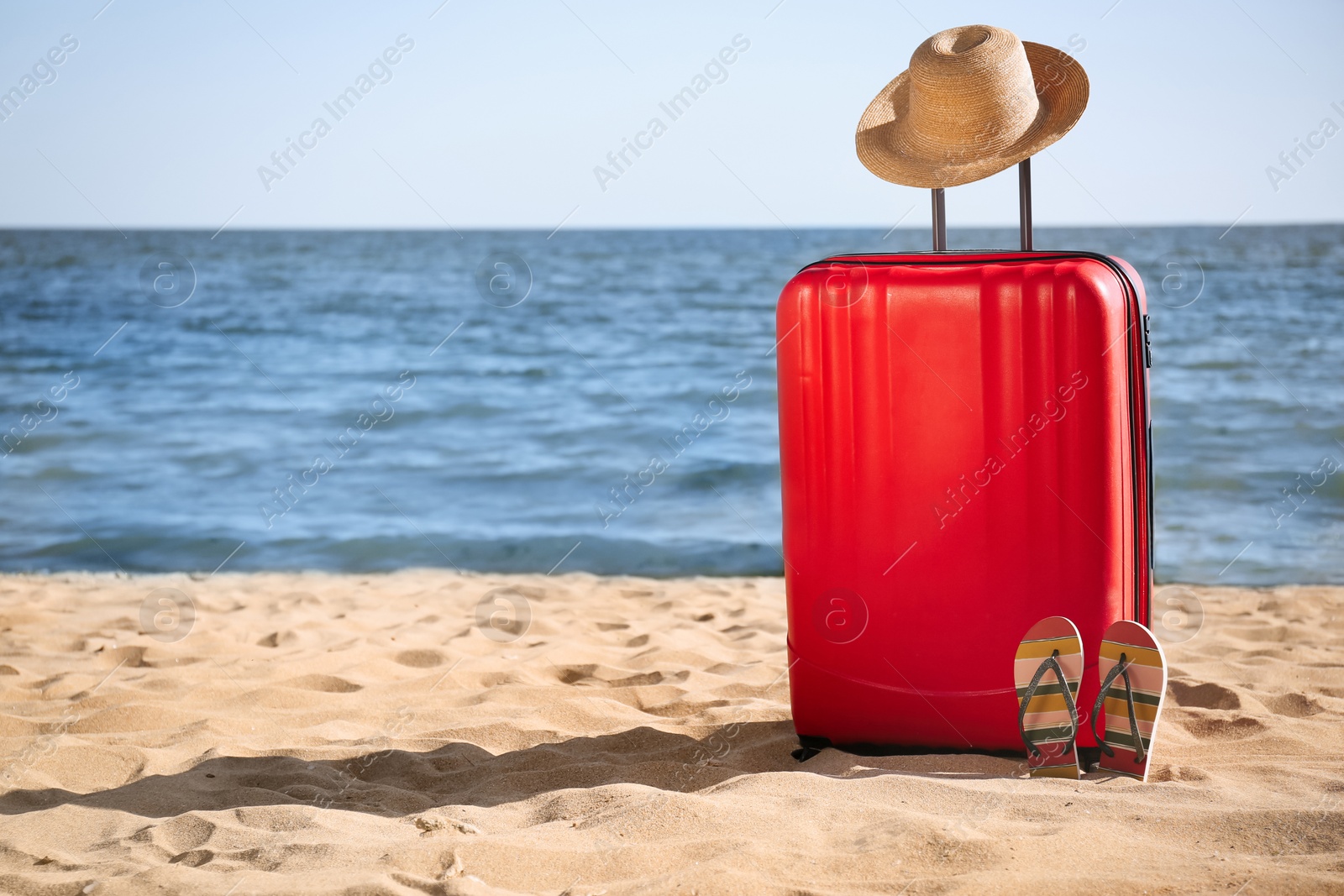 Photo of Bright suitcase, hat and flip flops on sand near sea, space for text. Beach accessories