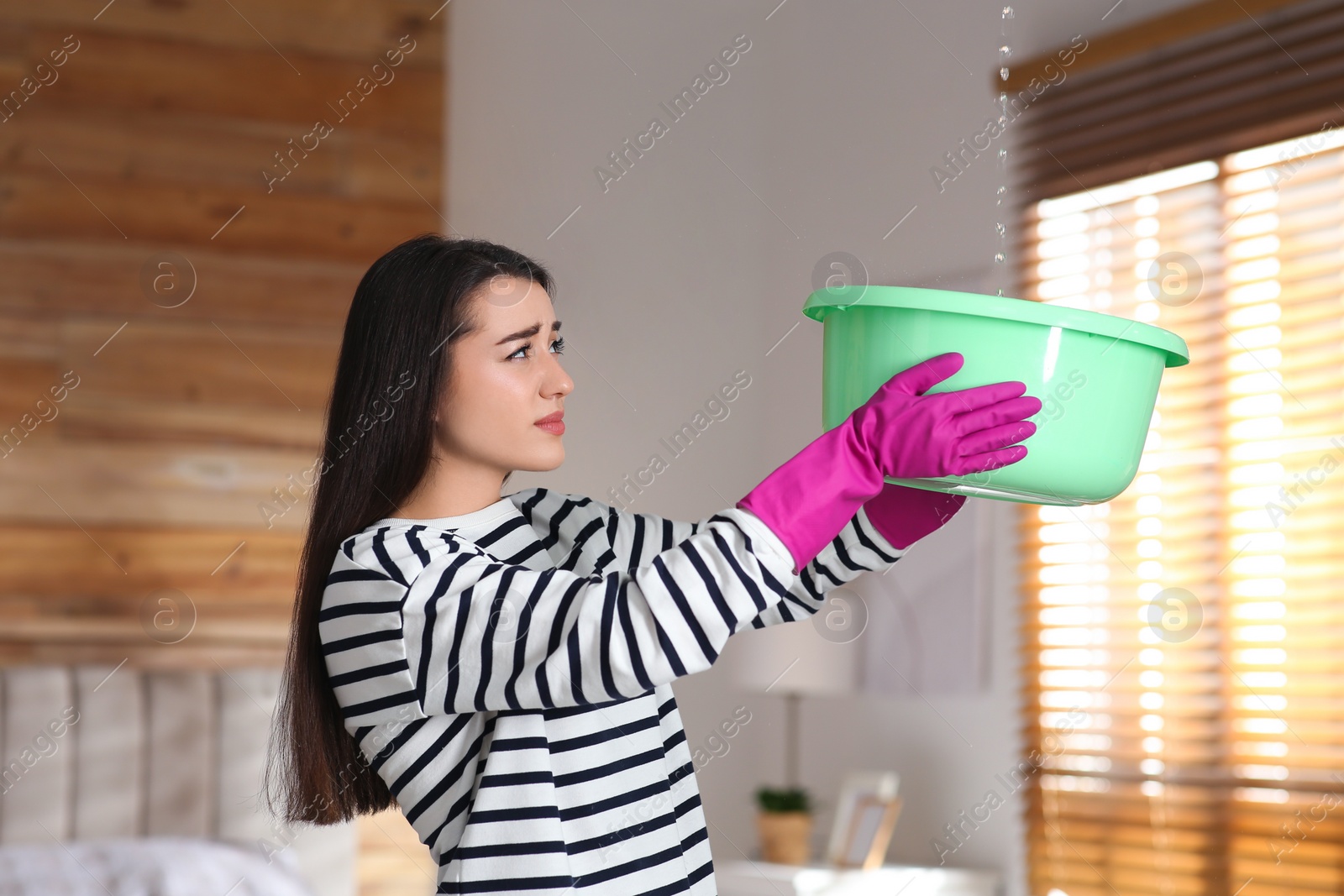 Photo of Young woman collecting leaking water from ceiling at home. Time to call roof repair service