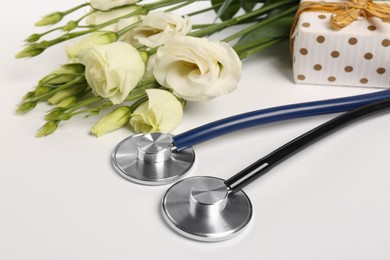 Photo of Stethoscopes, gift box and eustoma flowers on white background, closeup. Happy Doctor's Day
