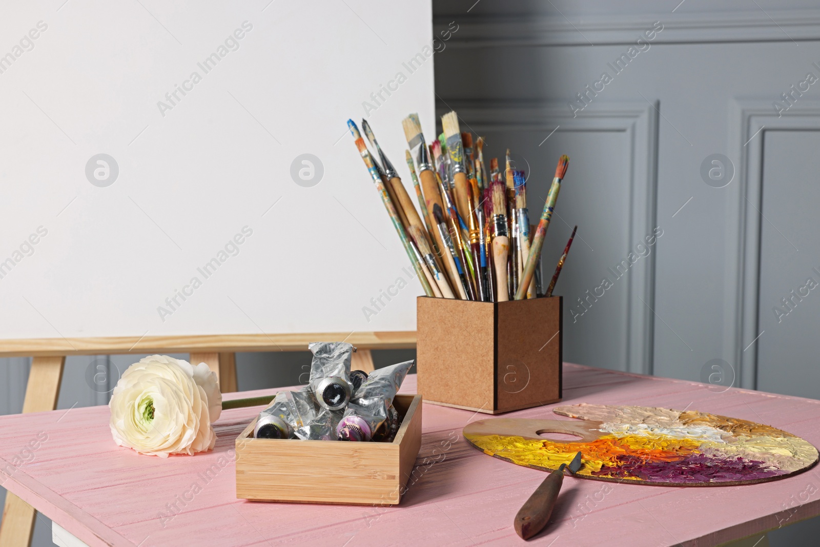 Photo of Easel with blank canvas and different art supplies on wooden table near grey wall