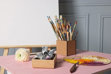 Easel with blank canvas and different art supplies on wooden table near grey wall