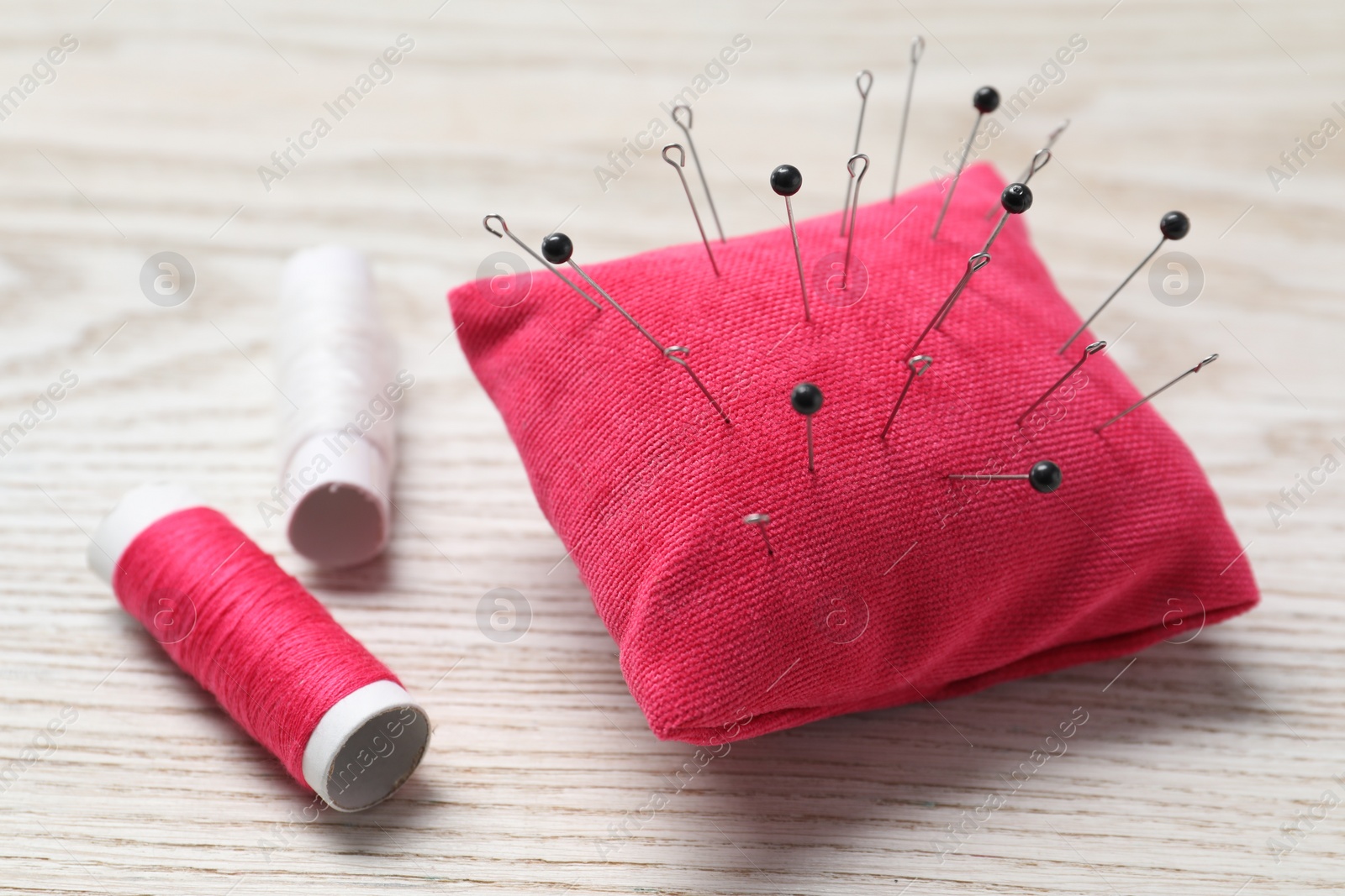 Photo of Red pincushion with sewing pins and spools of threads on light wooden table, closeup