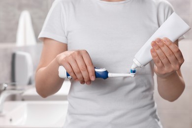Photo of Woman squeezing toothpaste from tube onto electric toothbrush in bathroom, closeup