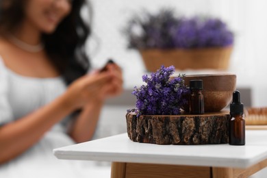 Photo of Woman sitting at table indoors, focus on stump with bottles of essential oil and flowers