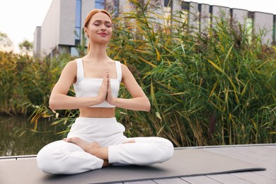 Beautiful young woman practicing Padmasana on yoga mat outdoors, low angle view. Lotus pose