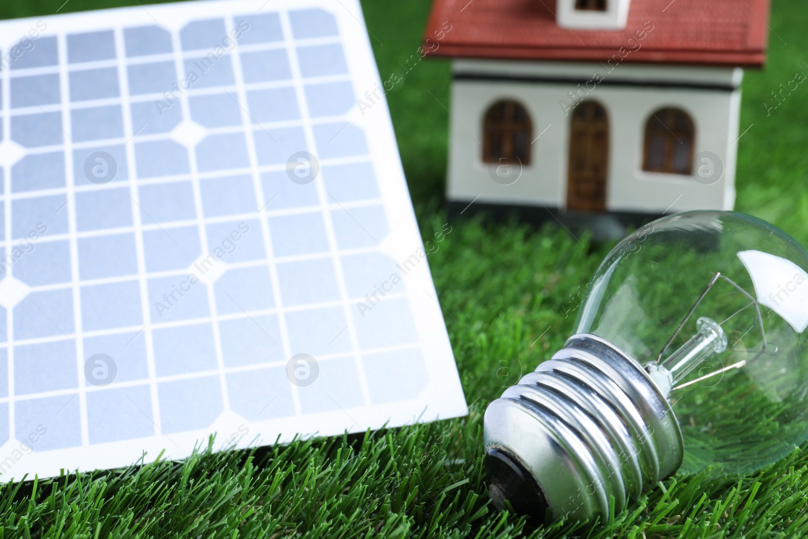 Photo of Solar panel, light bulb and house model on green grass, closeup