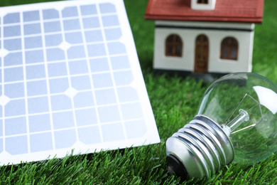 Photo of Solar panel, light bulb and house model on green grass, closeup