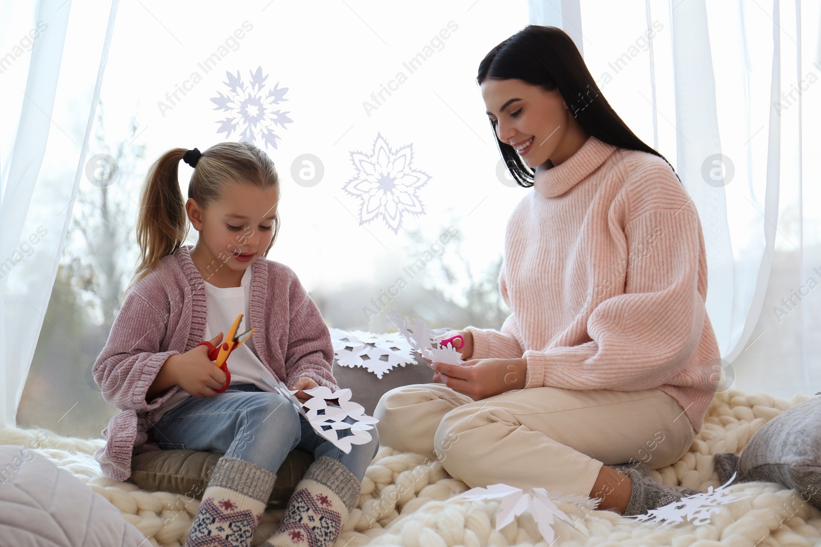 Photo of Mother and daughter making paper snowflakes near window at home