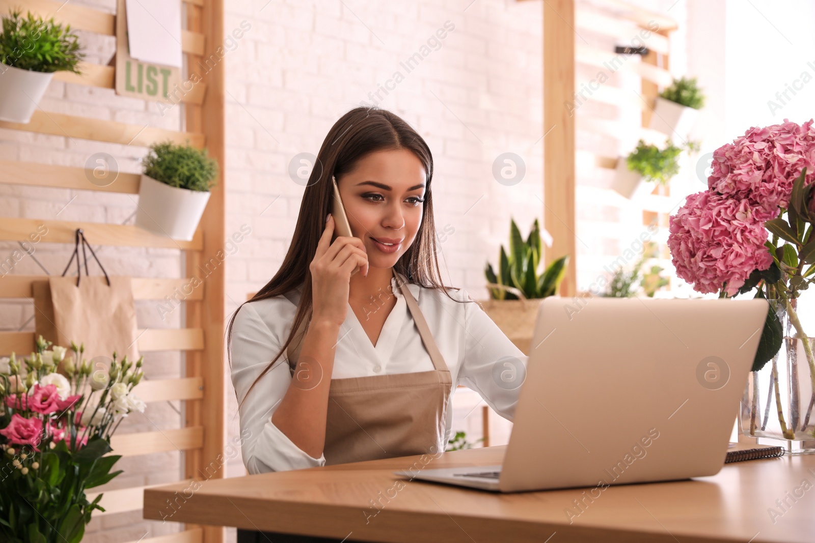 Photo of Florist talking on smartphone near laptop in workshop