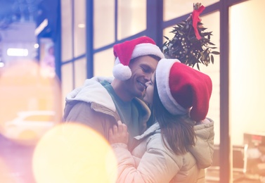 Happy couple in Santa hats kissing under mistletoe bunch outdoors, bokeh effect