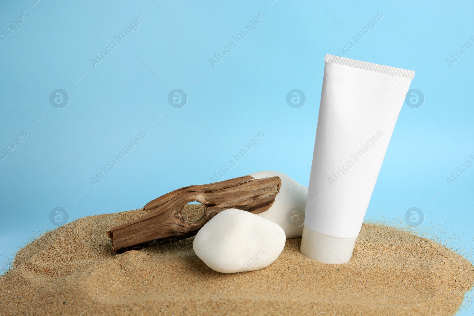 Photo of Tube of cream, stones and bark on sand against light blue background. Cosmetic product