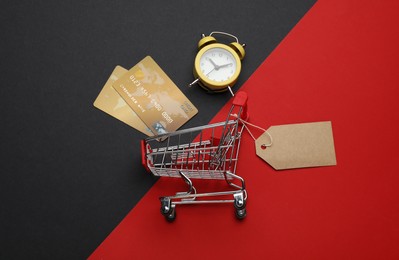 Photo of Shopping cart with price tag, credit cards and alarm clock on color background, flat lay