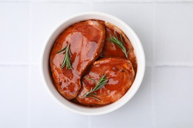 Raw marinated meat and rosemary in bowl on white tiled table, top view