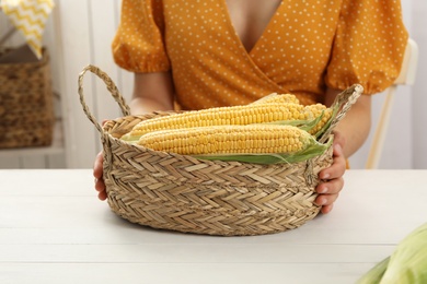 Woman with basket of corn cobs at white wooden table, closeup