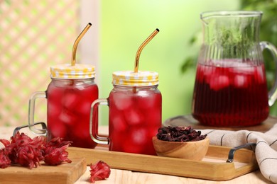 Refreshing hibiscus tea with ice cubes and roselle flowers on wooden table against blurred green background