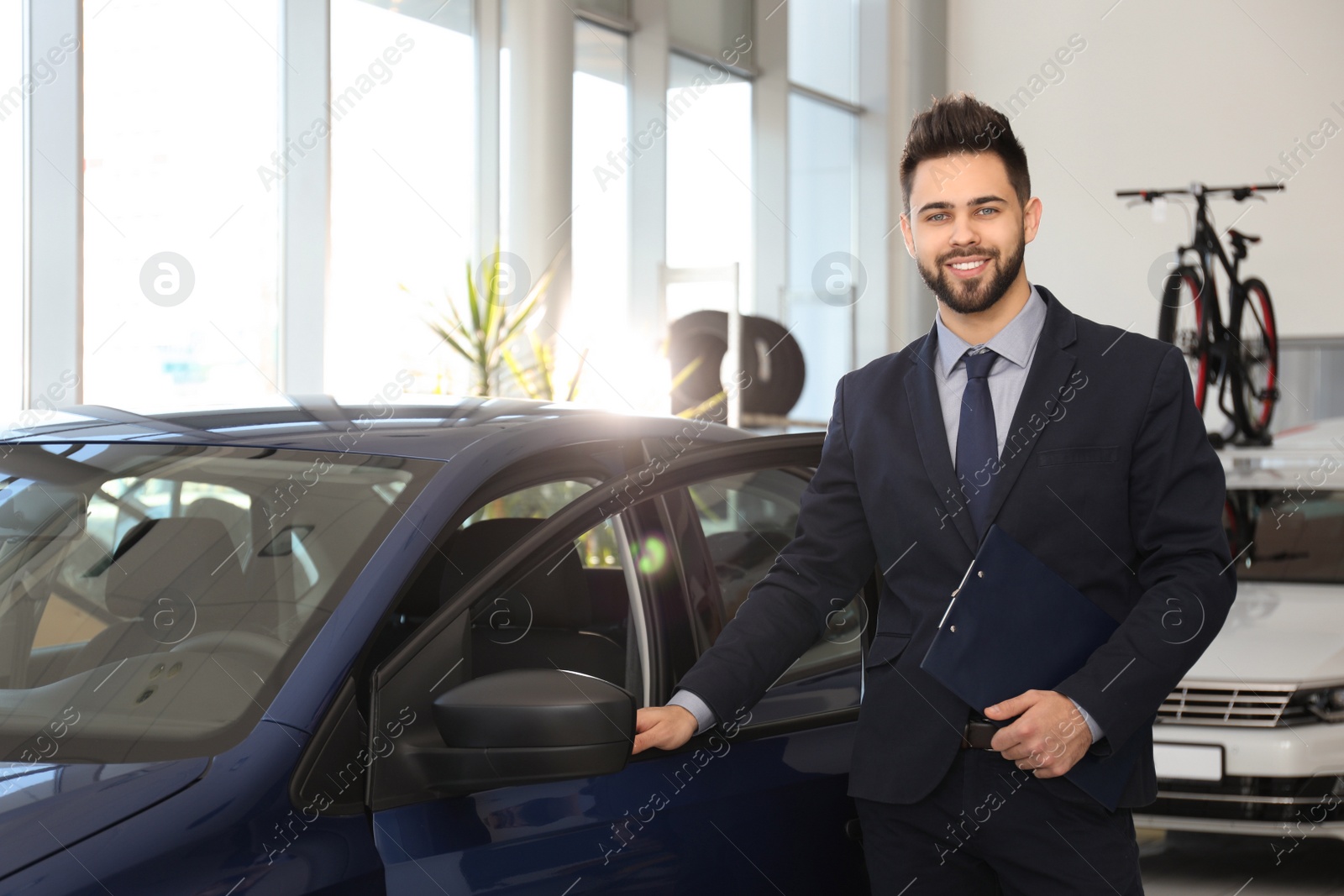 Photo of Young salesman with clipboard near car in dealership