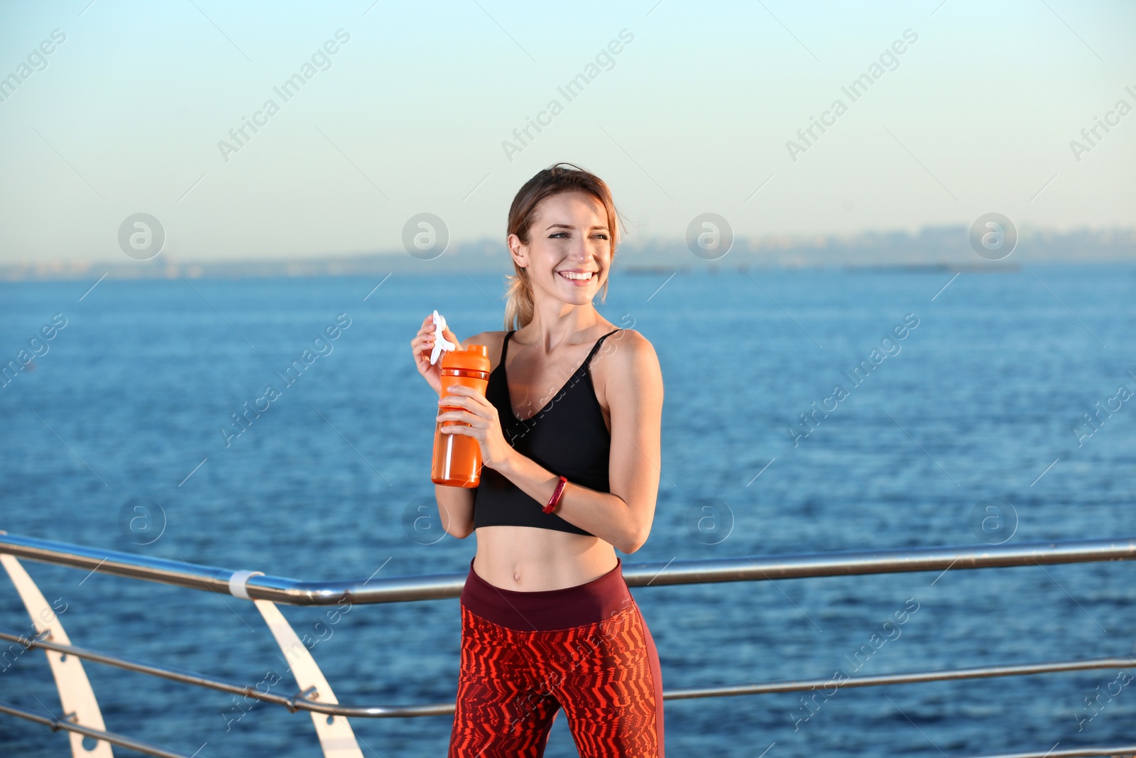 Photo of Young woman holding bottle with water on pier. Morning workout