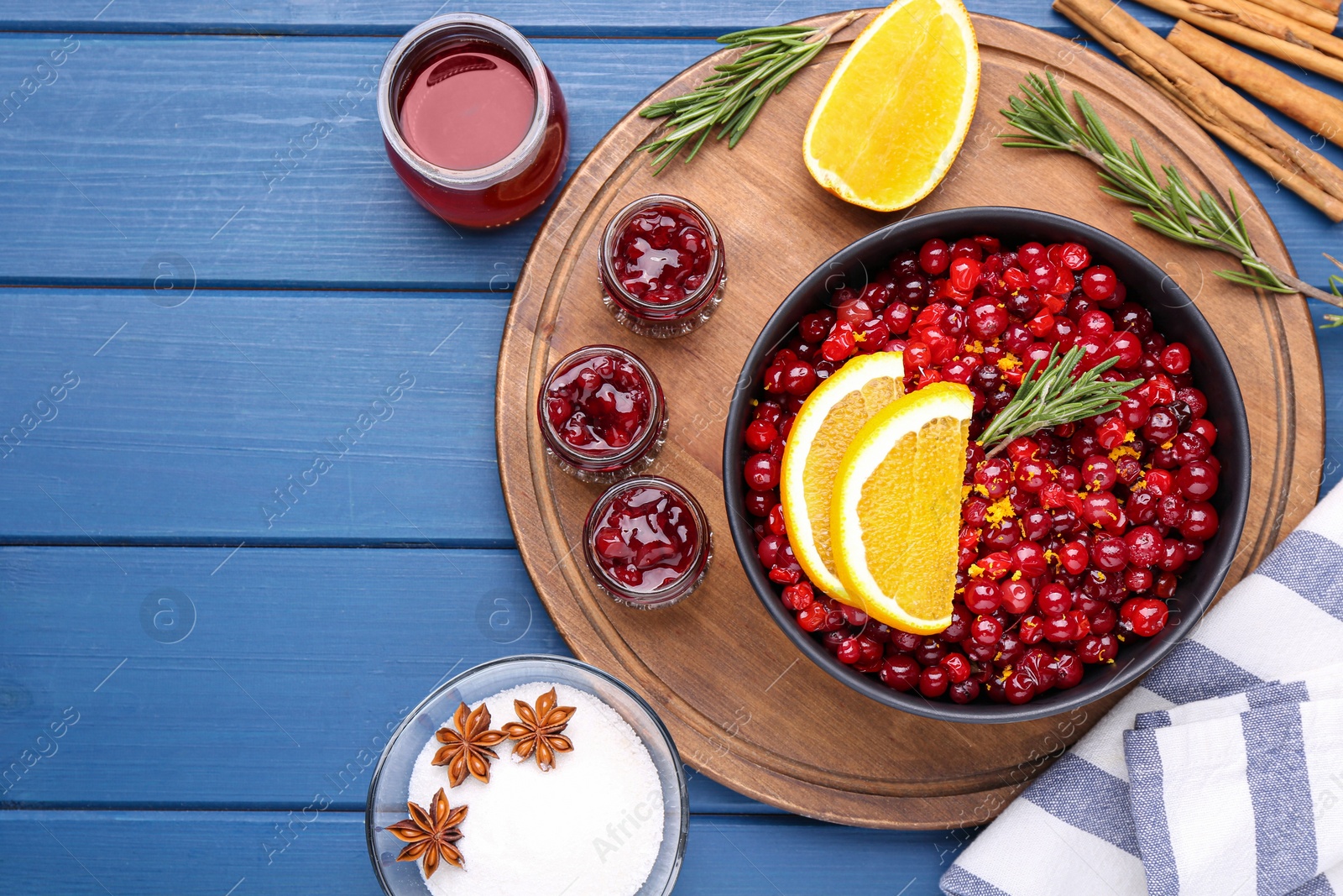 Photo of Cranberries in bowl, jars with sauce and ingredients on blue wooden table, flat lay. Space for text