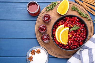 Cranberries in bowl, jars with sauce and ingredients on blue wooden table, flat lay. Space for text