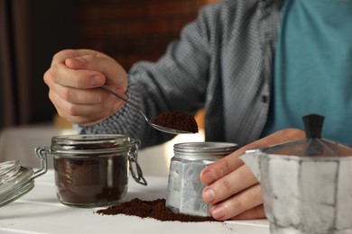 Man putting ground coffee into moka pot at white wooden table indoors, closeup